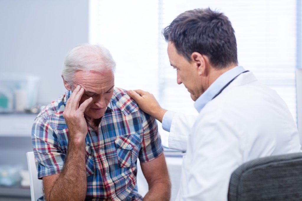 An older man looking upset as a doctor consoles him in a clinic.