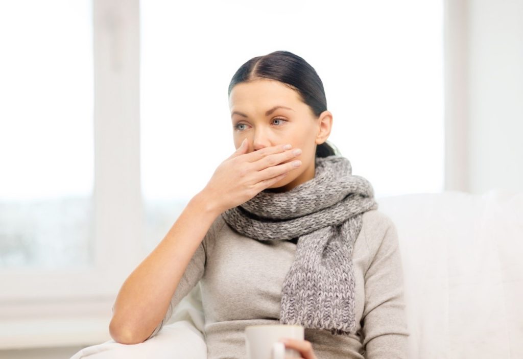 A sick woman holding a cup of tea about to sneeze.