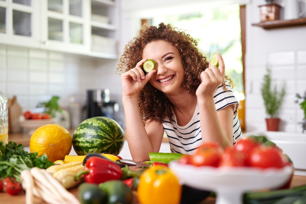 A smiling woman in a kitchen where she is surrounded by fruits and vegetables playfully holds a cucumber slice to her eye.
