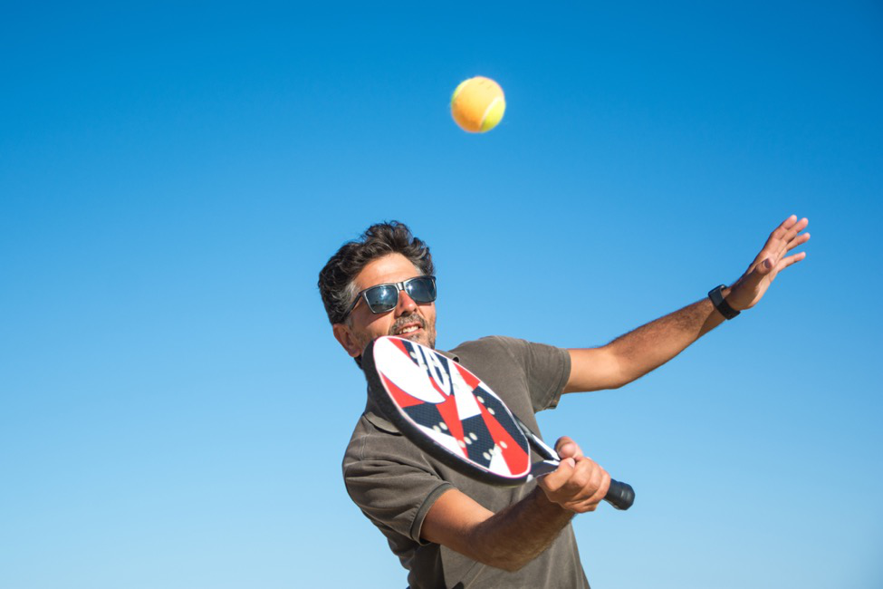 Man in sunglasses and gray shirt hitting a tennis ball with a red and blue paddle.