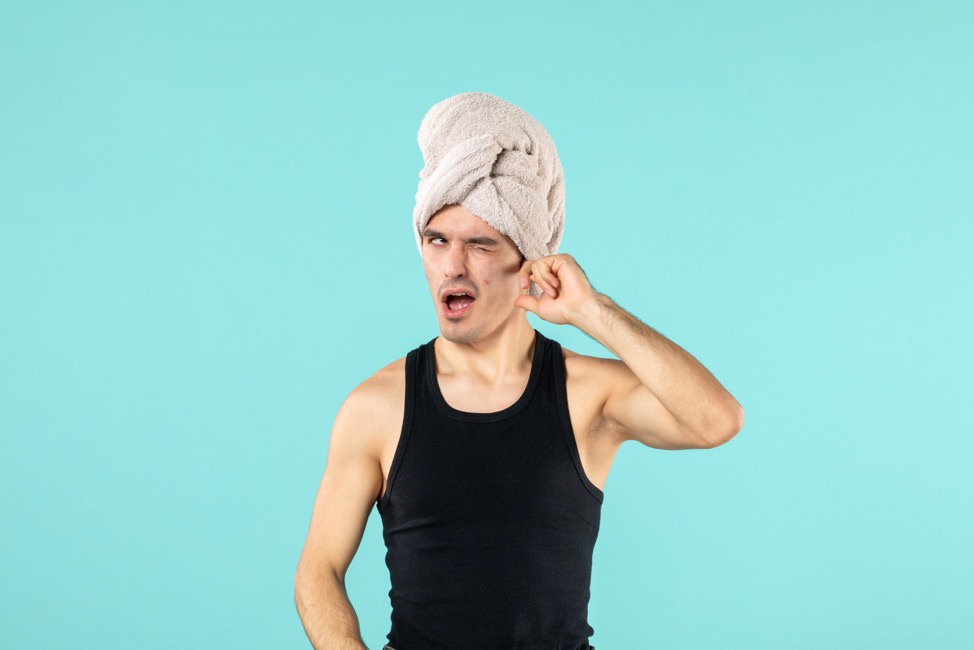 A man with swimmer’s ear holding a finger in his ear as he wears a towel on his head.