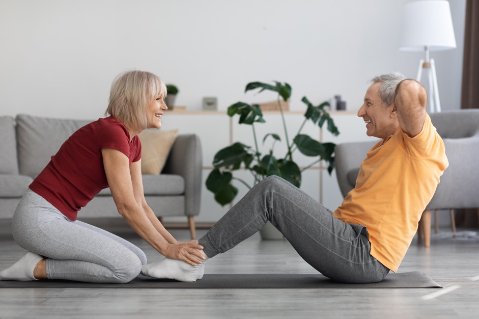 an older man does sit-ups while an older woman holds his ankles down.