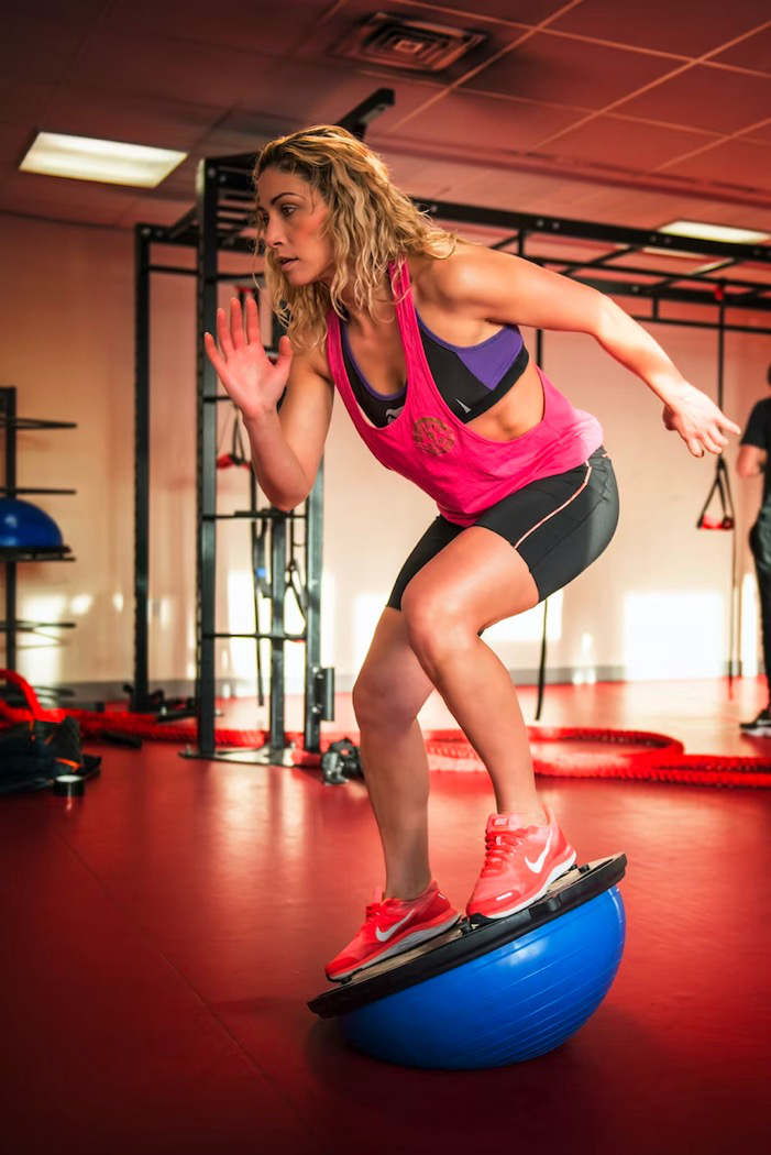 A woman stands on a Bosu ball while balancing during a workout.