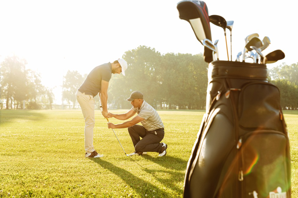 A golf instructor kneels before a player to help him adjust his swing.