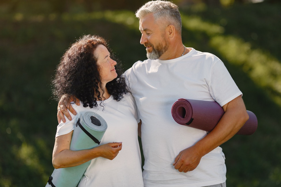 A man and a woman wearing white shirts, holding yoga mats while looking at each other.