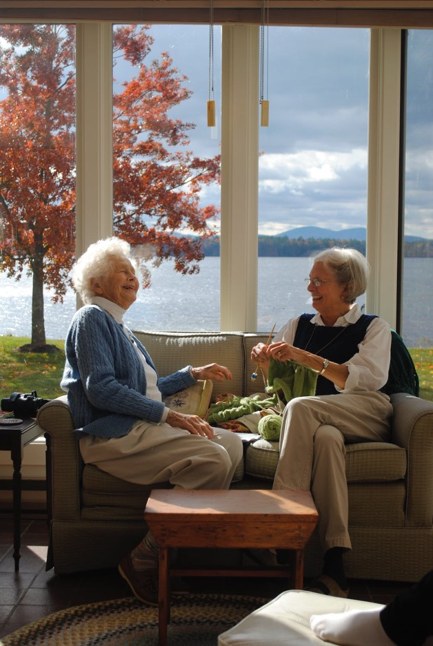 Two older women talking and laughing while sitting comfortably on a sofa overlooking a lake