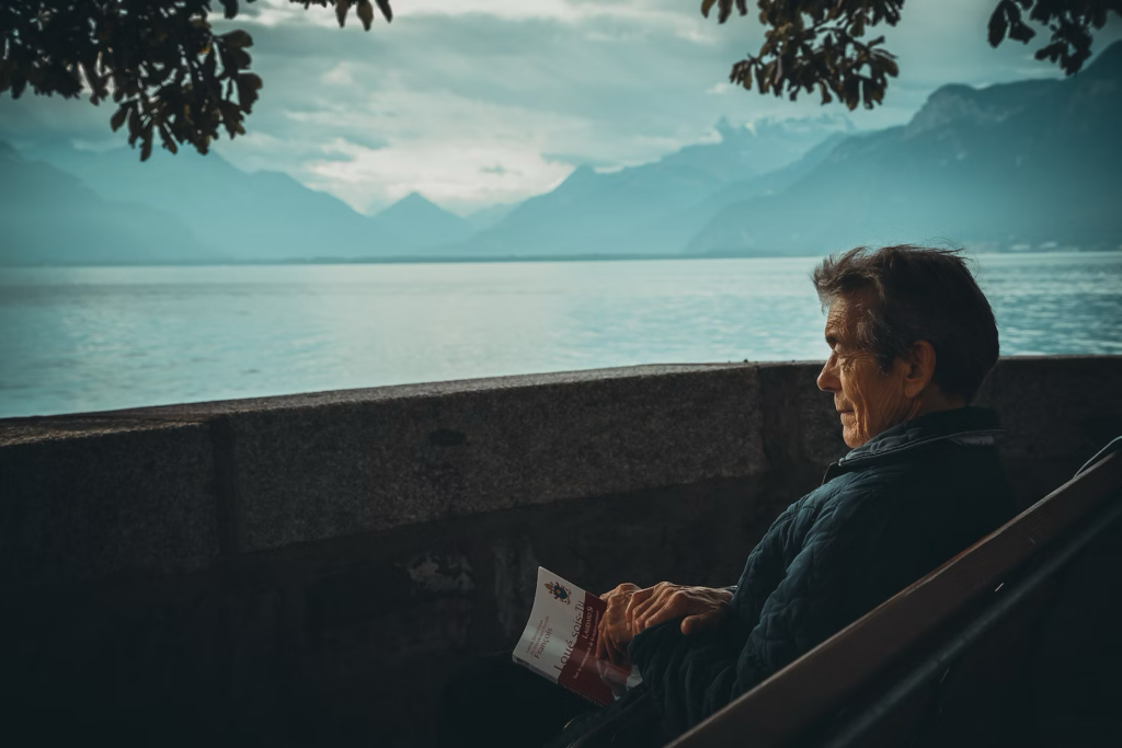 An elderly man reading a book while sitting by the sea.
