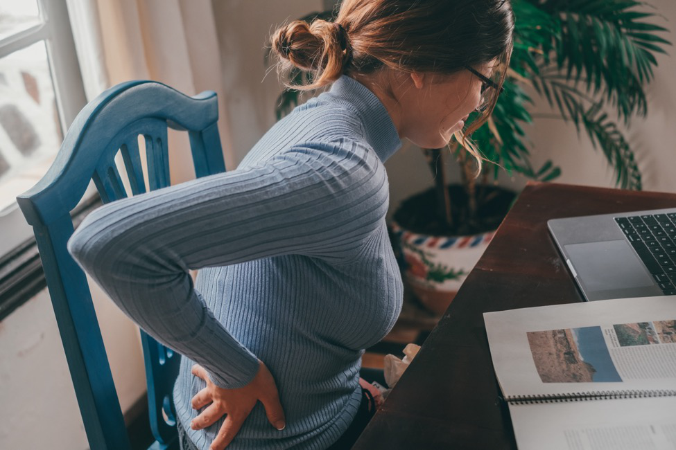 A woman sits in a chair while holding her lower back as though she is experiencing back pain.