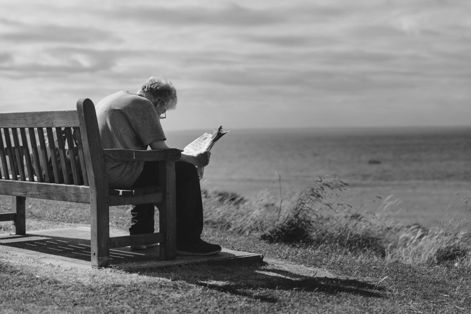 An older man reading a newspaper on a bench overlooking a vast field