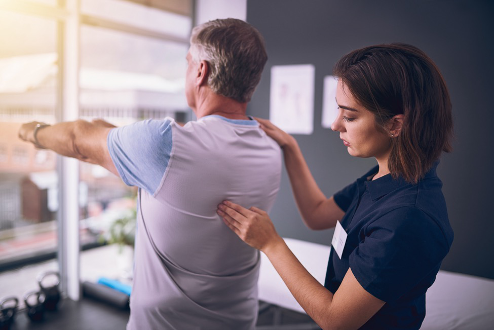 A man stands with his arms forward as a physiotherapist provides physical therapy for his back pain.