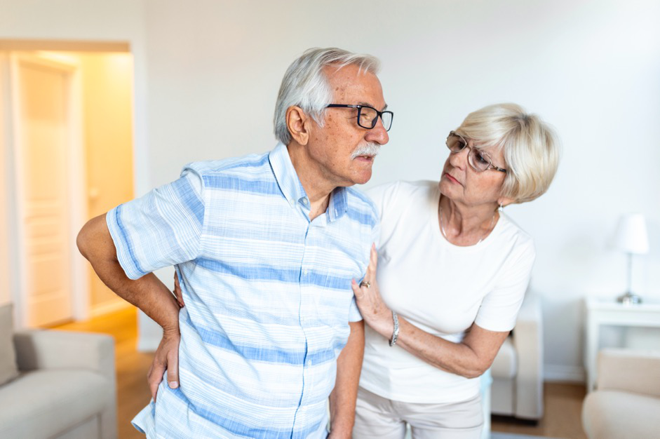 A man holding his lower back in pain while his concerned partner looks on in a home setting.