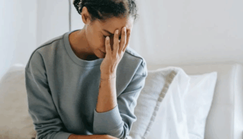 A woman in her late 30s sitting on the edge of a bed with her hand on her face, appearing overwhelmed by symptoms.