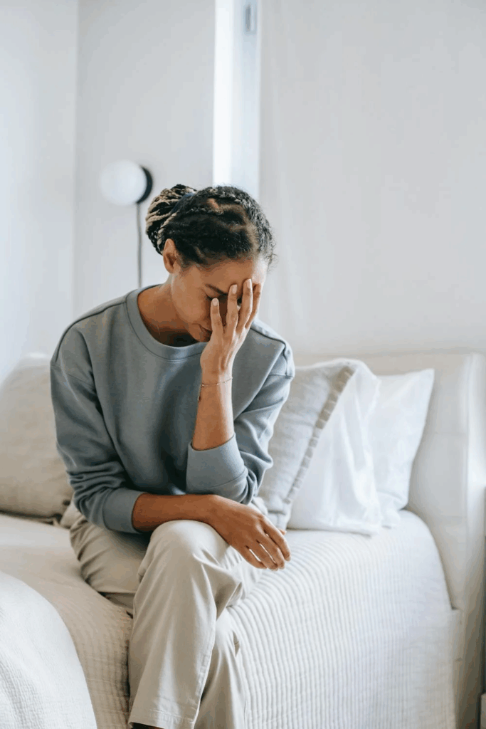 A woman in her late 30s sitting on the edge of a bed with her hand on her face, appearing overwhelmed by symptoms.