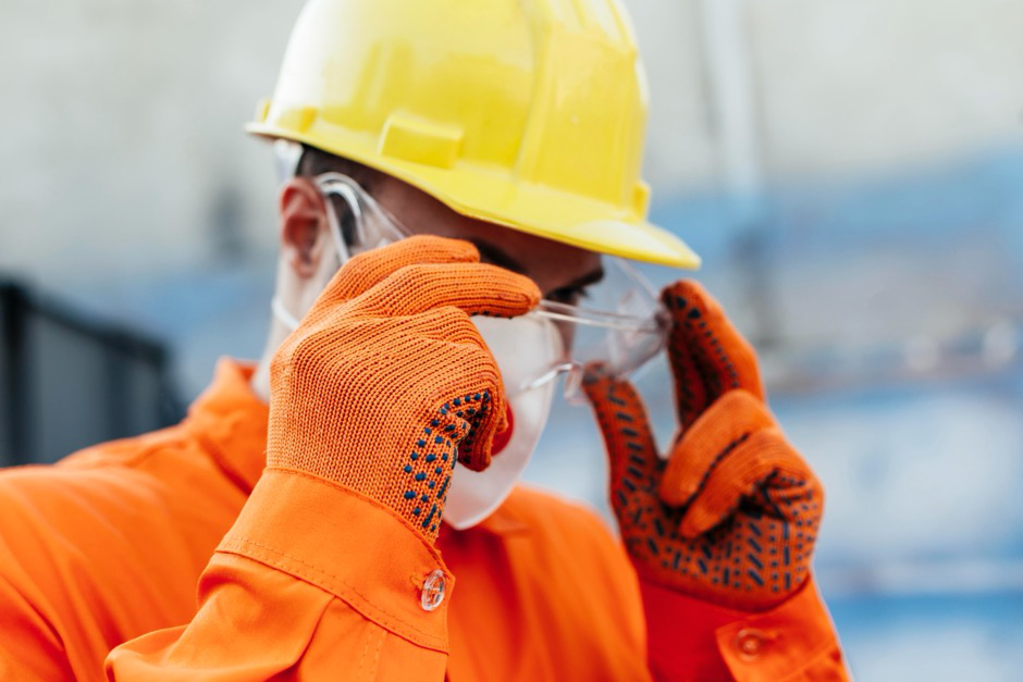 A construction worker on a site wearing a hard hat, gloves, mask, and safety glasses.