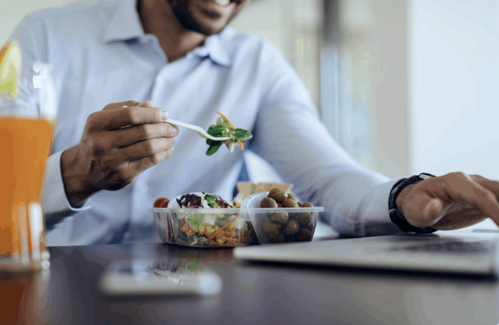 A young man smiling and eating a healthy salad at his desk.
